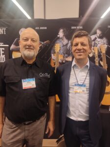 Photo of Miles Jackson and Alex Masso, standing in front of the Cole Clark stand at the NAMM Show with a row of acoustic guitars on display.